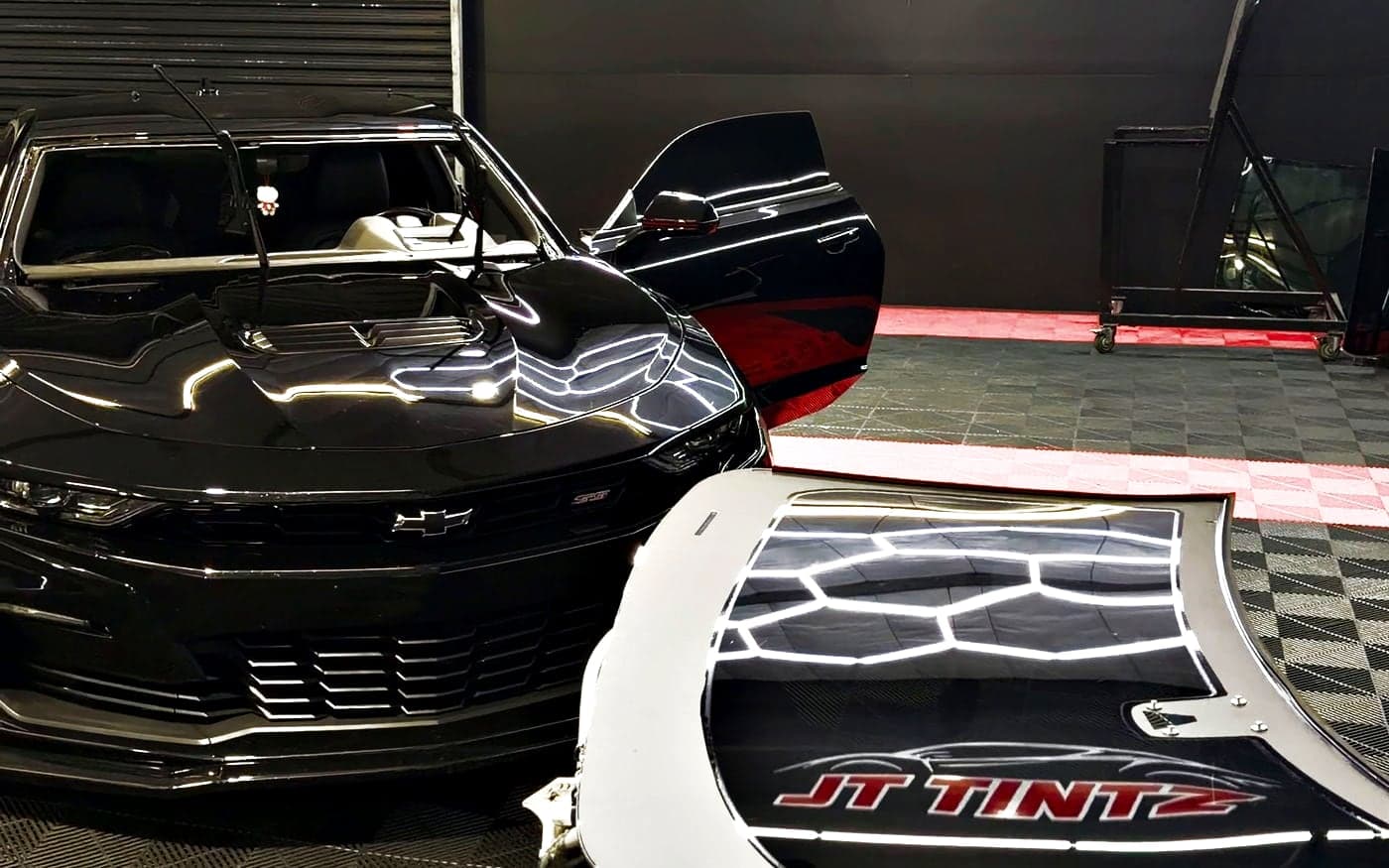 A black Camaro with its windshield removed, the glass resting on a stand in front of it reflecting the JT Tintz sign
