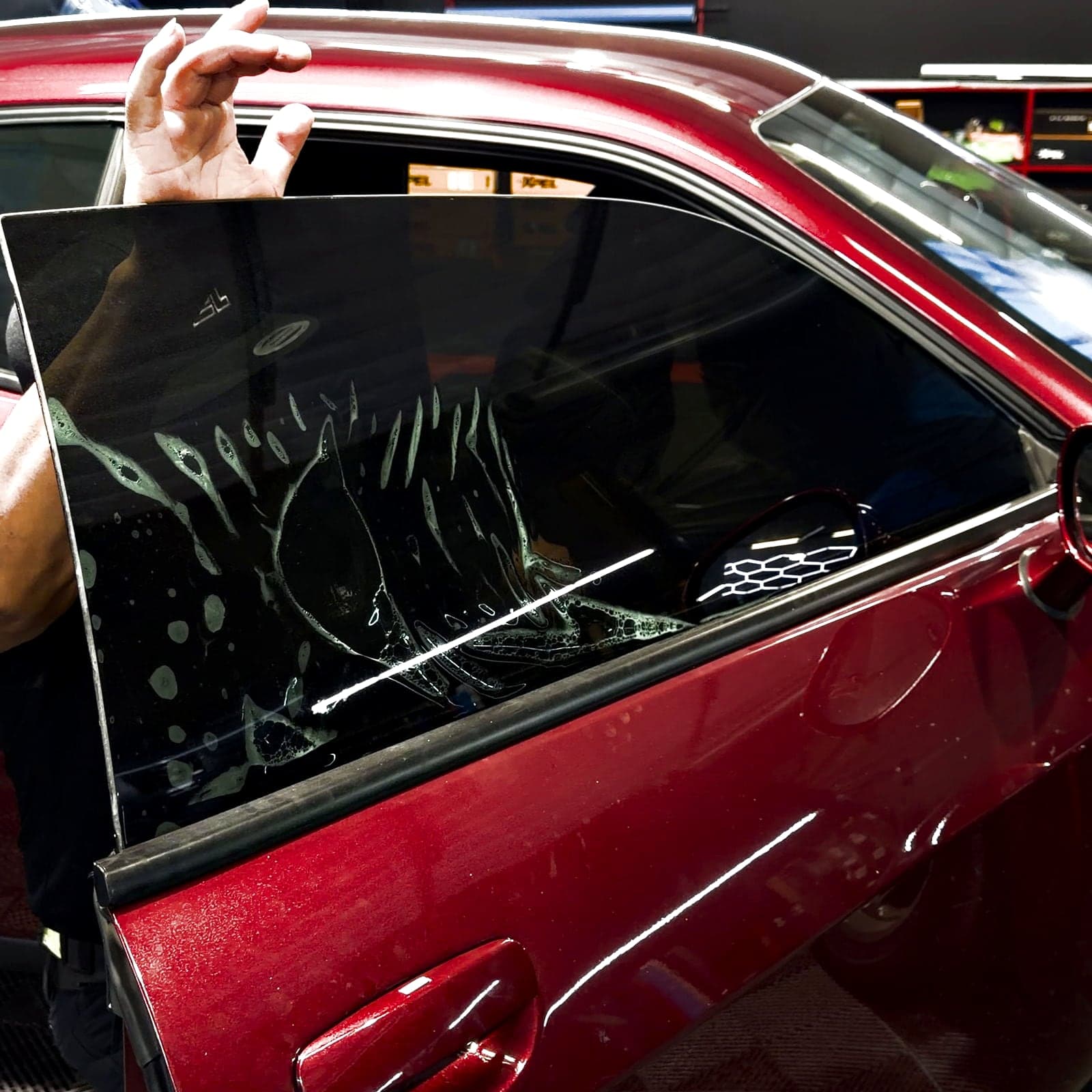 A sheet of tint film laid across the wet door glass of a dark red Challenger inside the JT Tintz bay